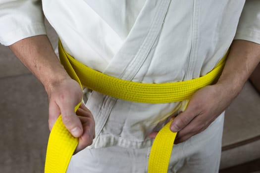 Man ties yellow Kyokushin karate belt on kimono around his waist, martial arts sports equipment before fight