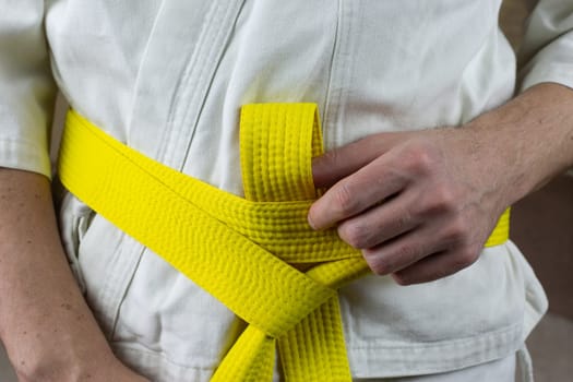 Male hands tighten yellow karate belt on kimono, close-up photo of Kyokushin karate equipment