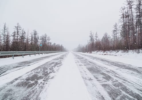 A long stretch of a deserted highway runs through a dense pine forest, blanketed in snow, under an overcast sky.