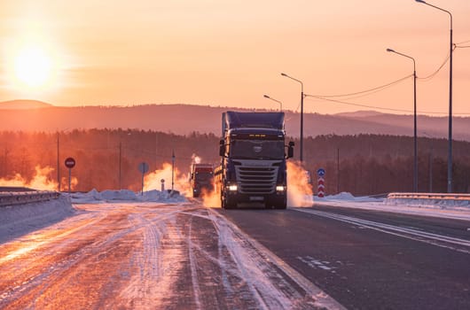 Two large trucks are driving along a snow-covered highway during a breathtaking winter sunrise.