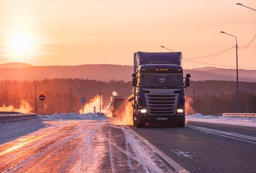 Two large trucks are driving along a snow-covered highway during a breathtaking winter sunrise.