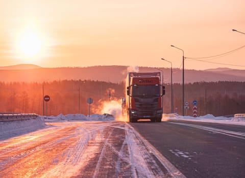 Two large trucks are driving along a snow-covered highway during a breathtaking winter sunrise.