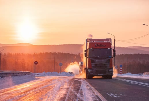 Two large trucks are driving along a snow-covered highway during a breathtaking winter sunrise.