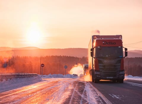 Two large trucks are driving along a snow-covered highway during a breathtaking winter sunrise.