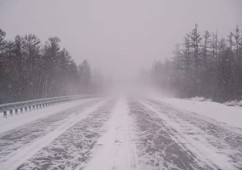 A remote highway runs through a dense forest, heavily covered in snow amid a fierce winter storm. Visibility is low, and the landscape is entirely white with streaks of snow crossing the road.