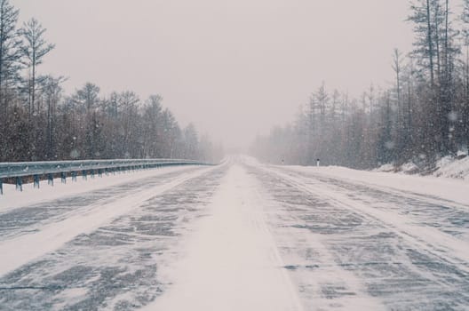 A remote highway runs through a dense forest, heavily covered in snow amid a fierce winter storm. Visibility is low, and the landscape is entirely white with streaks of snow crossing the road.