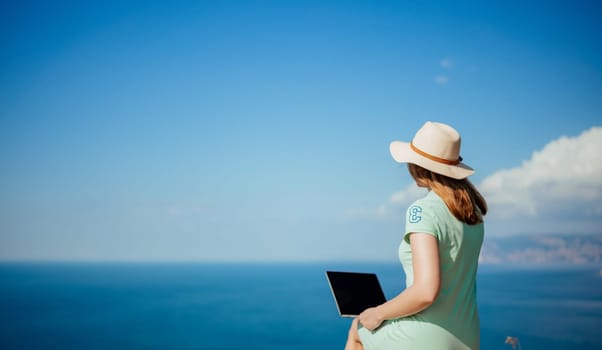 A woman is sitting on a rock by the ocean with a laptop in front of her. She is wearing a straw hat and a green dress. Concept of relaxation and leisure