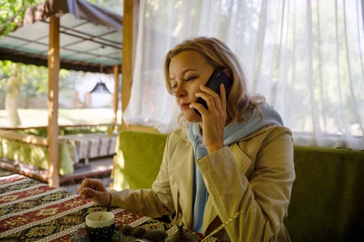 A woman is talking on her cell phone while sitting at a table. She is wearing a brown coat and a blue scarf