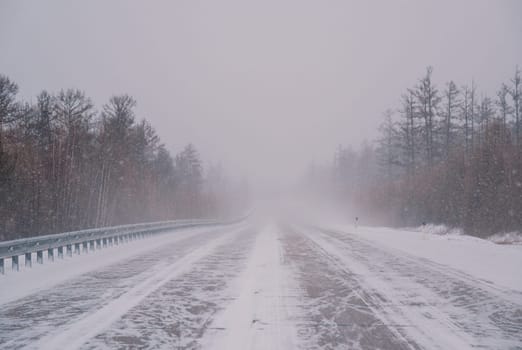 A remote highway runs through a dense forest, heavily covered in snow amid a fierce winter storm. Visibility is low, and the landscape is entirely white with streaks of snow crossing the road.