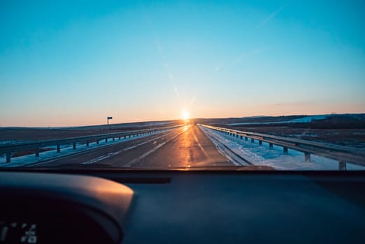 A scenic sunset is visible over a long, straight highway with snowy surroundings, seen from the perspective of the cars dashboard, giving a feeling of a road trip.