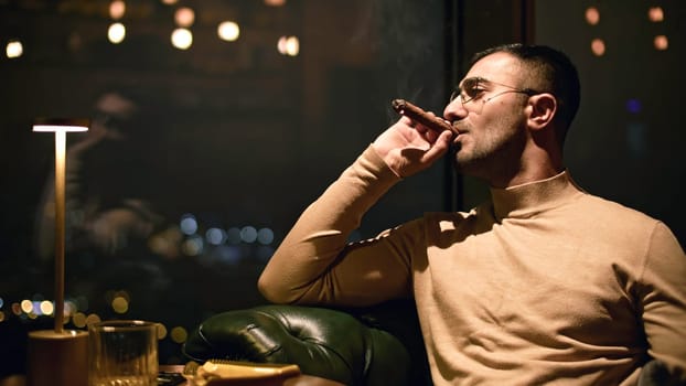 Businessman smokes cigar in restaurant. Media. Man smokes cigar in luxury restaurant at night. Elegant man smokes cigar in evening restaurant.