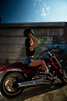 A brunette woman in denim shorts puts on a helmet while sitting on a red motorcycle. Vertical photo