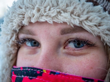 A close-up of a young womans face, showing her blue eyes and winter attire. She is bundled in a colorful scarf and a cozy knit hat, prepared for cold weather.