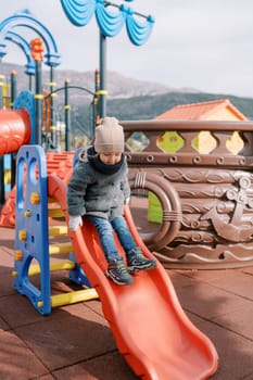 Little smiling girl in mittens slides down a slide on the playground. High quality photo