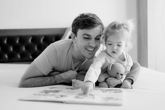 Little girl runs her finger along the lines of the book that her dad reads to her, lying on the bed. Black and white photo. High quality photo