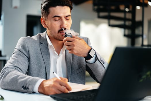 Young businessman smoking an electronic cigarette in his office while working close up