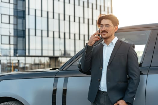 Young businessman in formal suit standing near luxury car and talking on the phone outdoors