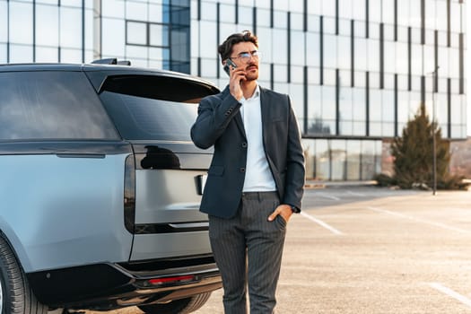 Young businessman in formal suit standing near luxury car and talking on the phone outdoors