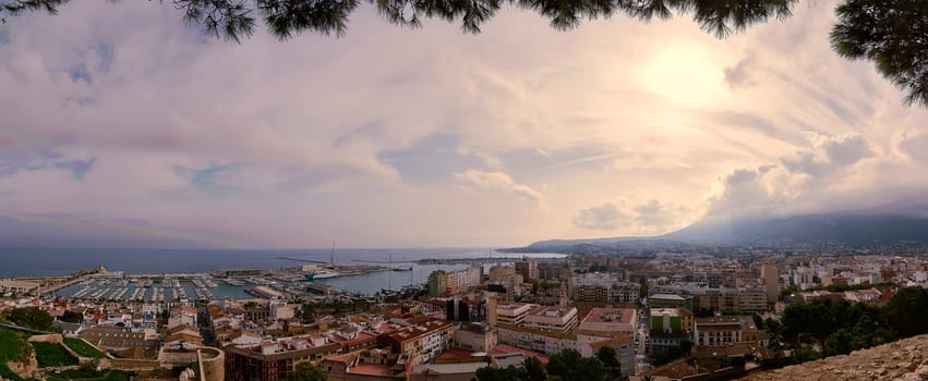 igh-resolution panoramic photograph showcasing the stunning coastal city of Denia, Spain, bathed in warm sunlight and adorned with fluffy clouds against a clear blue sky.