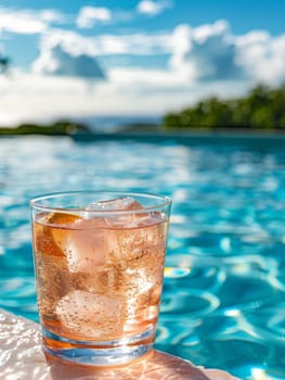 A glass of pink drink with ice cubes in it is sitting on a ledge by a pool. The drink is a refreshing beverage to enjoy on a hot day by the water