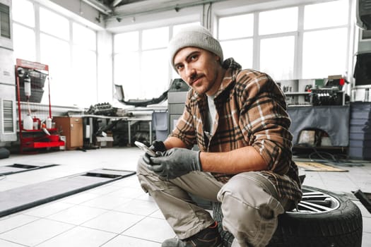 Car service worker sitting on a wheel and resting, using mobile phone close up