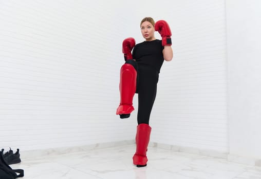 European blonde young kickboxing woman in black activewear and red kickboxing gloves, isolated on white background performing a martial arts kick. Sport exercise, fitness and cardio workout concept