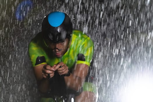 A triathlete braving the rain as he cycles through the night, preparing himself for the upcoming marathon. The blurred raindrops in the foreground and the dark, moody atmosphere in the background add to the sense of determination and grit shown by the athlete