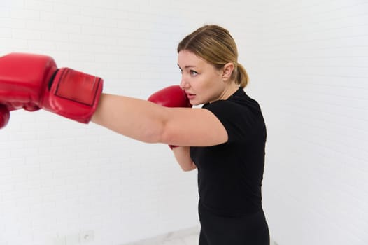 Side portrait of European woman 40s, female fighter boxer exercising with boxing gloves, punching forward, isolated over white wall background. People. Sport. Active healthy lifestyle concept