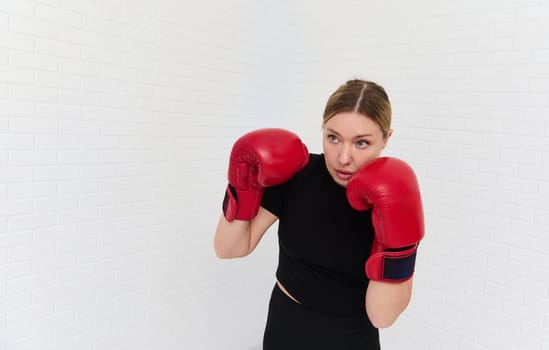 Confident determined young blond woman fighter, female boxer exercising with boxing gloves, punching forward, isolated over white bricks wall background. Martial art and sport concept. Copy ad space