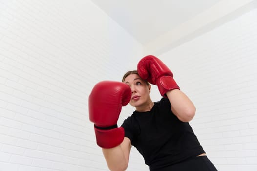 View from the bottom of Caucasian woman 40s, female fighter boxer exercising with boxing gloves, punching forward, isolated over white wall background. People. Sport. Active healthy lifestyle concept