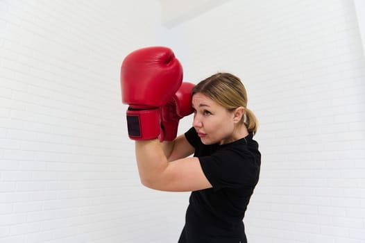 Portrait of a determined young female boxer in black sportswear and red boxing gloves, exercising, practicing box isolated over white background. Copy advertising space. People, sport and martial art