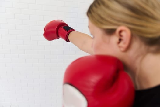 Close-up rear view of young blond woman fighter, determined boxer exercising with boxing gloves, punching forward, isolated over white bricks wall background. Martial art and sport concept. Copy space