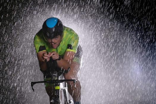 A triathlete braving the rain as he cycles through the night, preparing himself for the upcoming marathon. The blurred raindrops in the foreground and the dark, moody atmosphere in the background add to the sense of determination and grit shown by the athlete