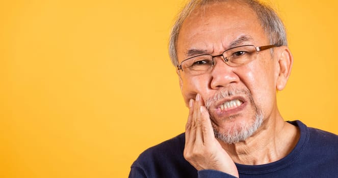 Dental pain. Asian unhappy elder man problems with gum pain studio shot isolated on yellow background, Portrait senior old man sad hand touching cheek suffering from toothache, dental healthcare