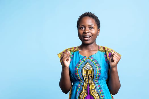 Charming girl in brightly colored attire looks startled when she sees something in studio against blue background. A girl of African American ethnicity dressed in ethnic traditional outfit.
