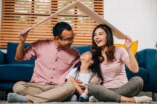 In their new home a joyful family holds a cardboard roof symbolizing investment and safety as they sit on a couch. Asian parents and their daughter exude happiness and togetherness.