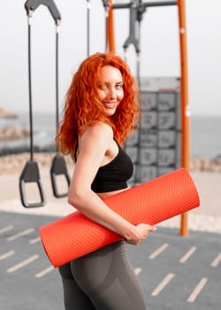 Smiling redhead sportswoman holding rolled yoga mat. Side view of fit strong female athlete in sportswear looking at camera while standing at outdoor gym on sunny day.