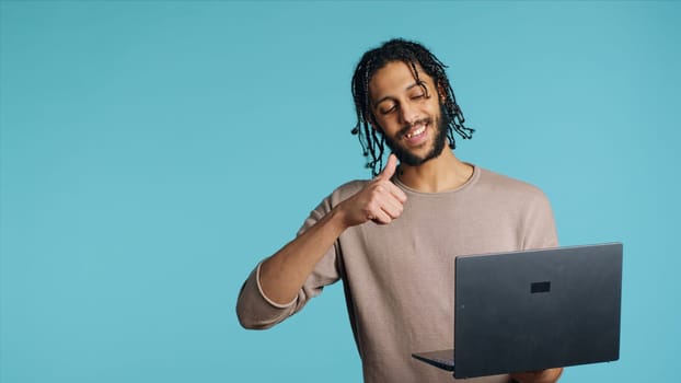 Middle Eastern man greeting friend over online videocall on laptop, doing friendly gestures. Happy person bonding with mate using notebook to hold videoconference, studio background, camera A