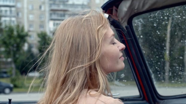 Young woman on passenger seat in a cabriolet car. Stock. Rain drops on car windshield and summer city on the background