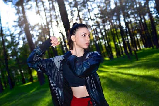 Young woman in a black sporty jacket doing arm exercises in a forest. Outdoor fitness.
