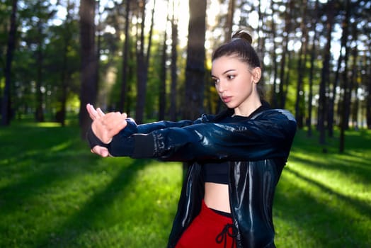 Young woman in a black sporty jacket doing arm exercises in a forest. Outdoor fitness.