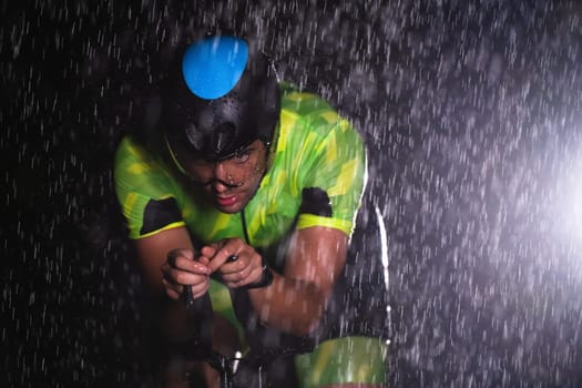 A triathlete braving the rain as he cycles through the night, preparing himself for the upcoming marathon. The blurred raindrops in the foreground and the dark, moody atmosphere in the background add to the sense of determination and grit shown by the athlete