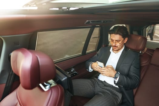 Young elegant businessman sitting on the back seat of a luxury car and using smartphone close up