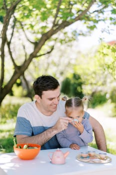 Dad treats his little daughter to tea from a toy cup while sitting at a table in the garden having tea party. High quality photo