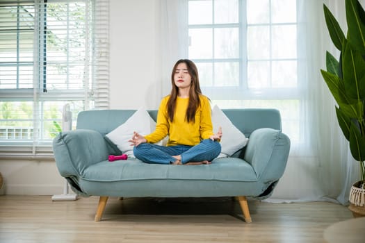 Asian young woman sits on sofa at home and chilling with couch lotus pose eyes closed, female practise yoga and meditation in lotus position, mindfulness lifestyle, happy relaxation leisure people