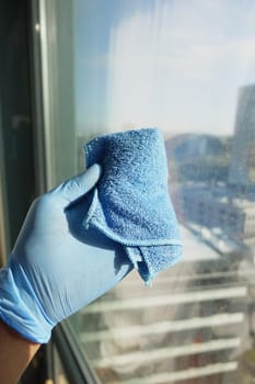 hand in blue glove cleaning window with green rag.