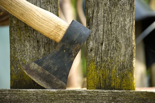 Close-up view of an axe, featuring a brown handle and dark grey blade, lodged firmly in an old, weathered wooden fence post with visible cracks