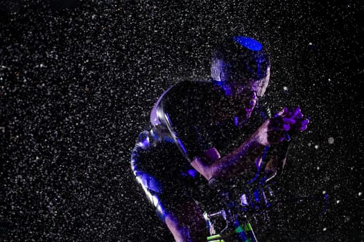 A triathlete braving the rain as he cycles through the night, preparing himself for the upcoming marathon. The blurred raindrops in the foreground and the dark, moody atmosphere in the background add to the sense of determination and grit shown by the athlete