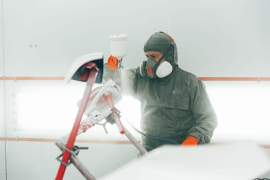 Auto mechanic worker wearing protective workwear spraying white paint on car part at workshop close up