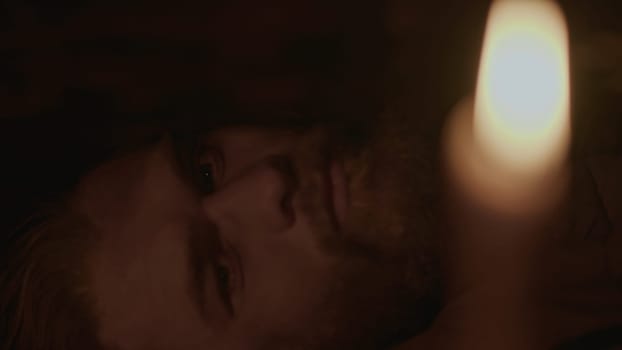 Close up of young bearded man closing eyes and falling asleep in a dark room with candlelight. Media. Pensive man lying and sleeping at night indoors